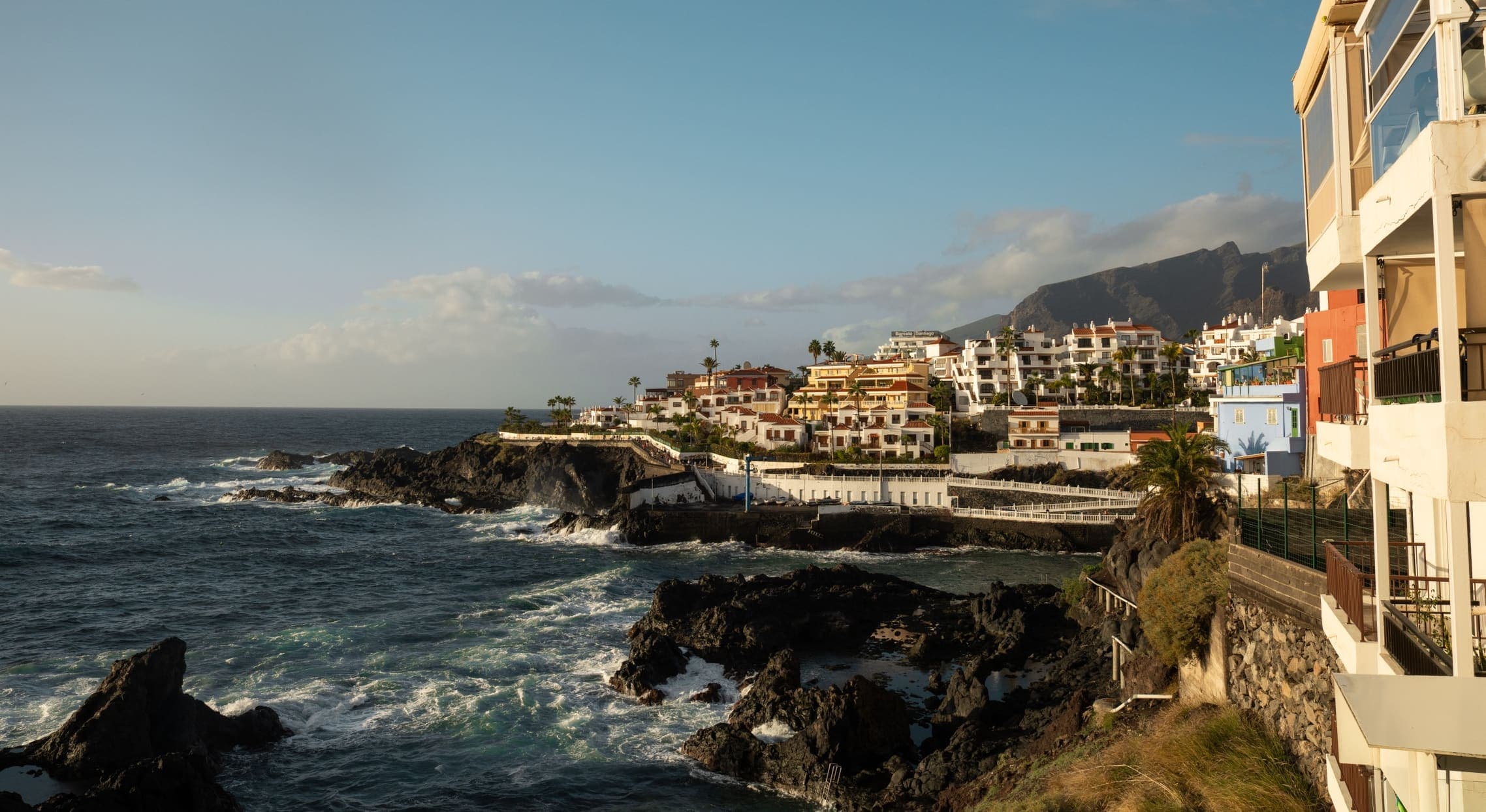 Sunlit homes along a rocky coastline, with mountains behind